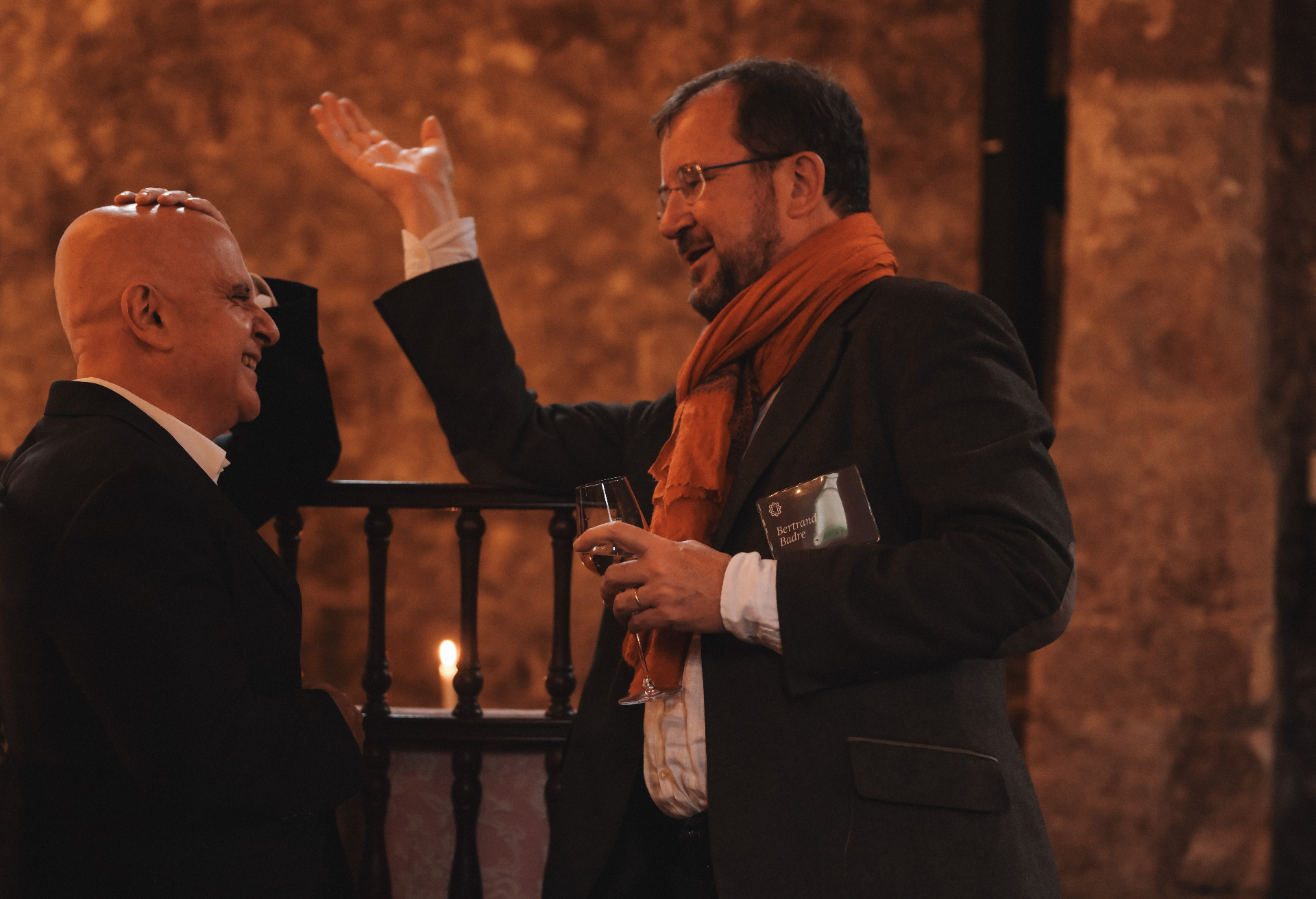 Two Odyssey guests sharing a warm exchange over wine in a candlelit stone hall.
